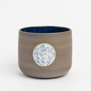 Op de afbeelding: Een kleine, grijze keramische beker met een blauwe binnenkant en een witte cirkel met blauwe stippen aan de zijkant.
