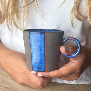 May include: A blue and grey ceramic mug with a unique handle. The mug has a blue and white glaze with a thin orange stripe.