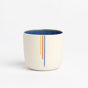 May include: A cream-coloured ceramic cup with a blue interior and three vertical stripes in red, yellow, and blue. The cup has a rounded shape and a simple, modern design. The cup is suitable for holding hot or cold beverages.