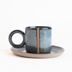 Pode incluir: Um conjunto de caneca e pires de cerâmica. A caneca é cilíndrica com uma alça grande e circular e uma faixa vertical laranja. A caneca e o pires são em tons de azul e cinza, com uma base de pires bege claro. O conjunto é projetado para bebidas.
