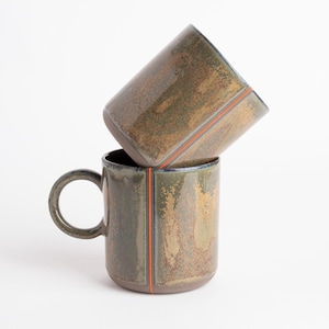 Pode incluir: Duas canecas de cerâmica em tons de verde e castanho, com uma faixa vertical laranja. Uma caneca está empilhada sobre a outra. As canecas têm uma capacidade de aproximadamente 350 ml. O design é moderno e as canecas têm uma pega.