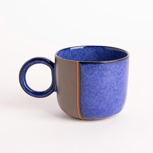 May include: A ceramic mug with a rounded body and a circular handle. The mug features a textured blue exterior, a dark gray side panel with an orange vertical stripe, and a dark blue interior. The mug is designed for hot or cold beverages.