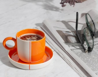 May include: A white and orange ceramic coffee cup with a white saucer. The cup is filled with coffee and there is a newspaper and glasses in the background.