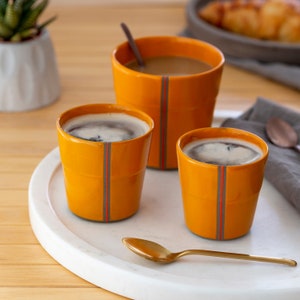 Puede incluir: Tres tazas de cerámica naranja con rayas grises, dos llenas de café y una de té, en una bandeja de mármol blanco con una cuchara de oro.