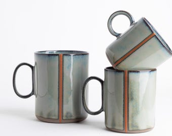 May include: Three ceramic mugs with a glossy, mottled light blue and gray glaze. Each mug features a vertical orange stripe running down the center and a dark blue rim. The mugs have a cylindrical shape with a single handle on the side.