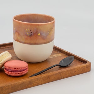 May include: A wooden tray with a pink and white ceramic mug with gold accents, a spoon, and two macarons. The macarons are pink and white.