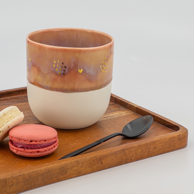 May include: A wooden tray with a pink and white ceramic mug with gold accents, a spoon, and two macarons. The macarons are pink and white.