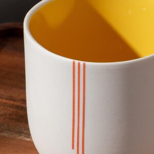 Peut inclure: Une tasse en c&eacute;ramique blanche avec un int&eacute;rieur jaune et trois fines lignes orange. La tasse est pos&eacute;e sur une surface en bois. Le texte "MODERN AESTHETIC" est sous la tasse. Le texte "SHOP NOW" et "@jasminsclayworks" sont sous le texte.