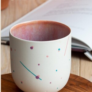 May include: A white ceramic mug with a pink, blue, and purple speckled design. The mug is sitting on a wooden surface. "SHOP NOW" is written in white text on a brown background.