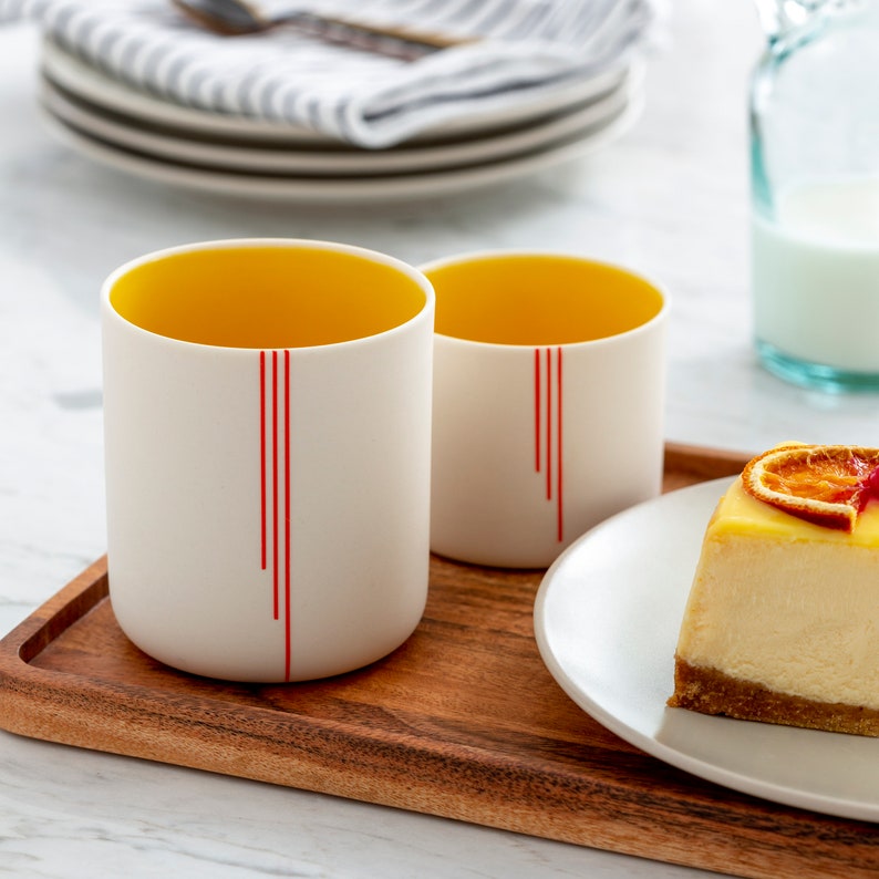 Peut inclure: Deux tasses en c&eacute;ramique blanche avec des int&eacute;rieurs jaunes et des lignes verticales rouges. Les tasses sont sur un plateau en bois avec une part de cheesecake sur une assiette blanche.