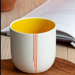 May include: A white ceramic mug with a yellow interior and orange stripes. The mug is sitting on a wooden surface. "SHOP NOW" is written on the surface.