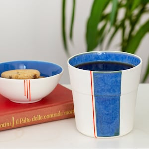 Op de afbeelding: Een witte keramische mok met een blauw en groen streepontwerp en een rode lijn. De mok is gevuld met koffie en staat op een wit oppervlak.