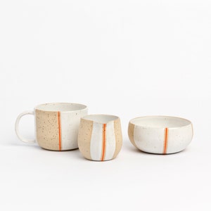 May include: A set of three ceramic mugs in a speckled white and beige glaze with a single orange stripe. The mugs are a mug, a creamer, and a bowl.