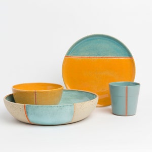 K&ouml;nnte beinhalten: Ein Set aus Keramikgeschirr in Blau, Gelb und Wei&szlig; mit orangefarbenen Akzenten. Das Set enth&auml;lt eine gro&szlig;e Sch&uuml;ssel, eine kleinere Sch&uuml;ssel, einen Teller und eine Tasse.