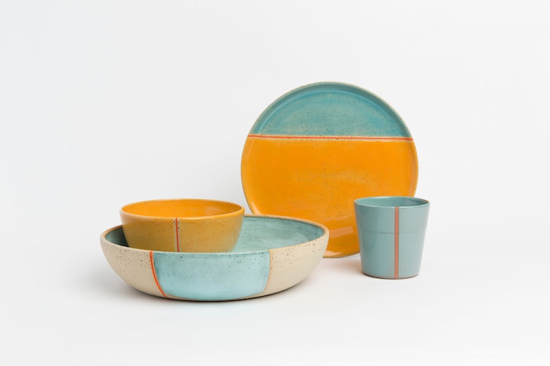 K&ouml;nnte beinhalten: Ein Set aus Keramikgeschirr in Blau, Gelb und Wei&szlig; mit orangefarbenen Akzenten. Das Set enth&auml;lt eine gro&szlig;e Sch&uuml;ssel, eine kleinere Sch&uuml;ssel, einen Teller und eine Tasse.