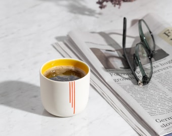 May include: A white ceramic mug with a yellow rim and orange stripes. The mug is filled with black coffee and is sitting on a white marble surface next to a stack of newspapers and a pair of glasses.