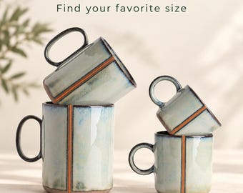 May include: Handmade stoneware mugs in a pale blue glaze with a vertical orange stripe and dark rim. The text reads: HANDMADE STONEWARE Everyday cups. Extraordinary rituals. Find your favorite size. Mugs sold individually. Explore the collection on Etsy. YASEMIN UGURLU ARTISAN CERAMICS.