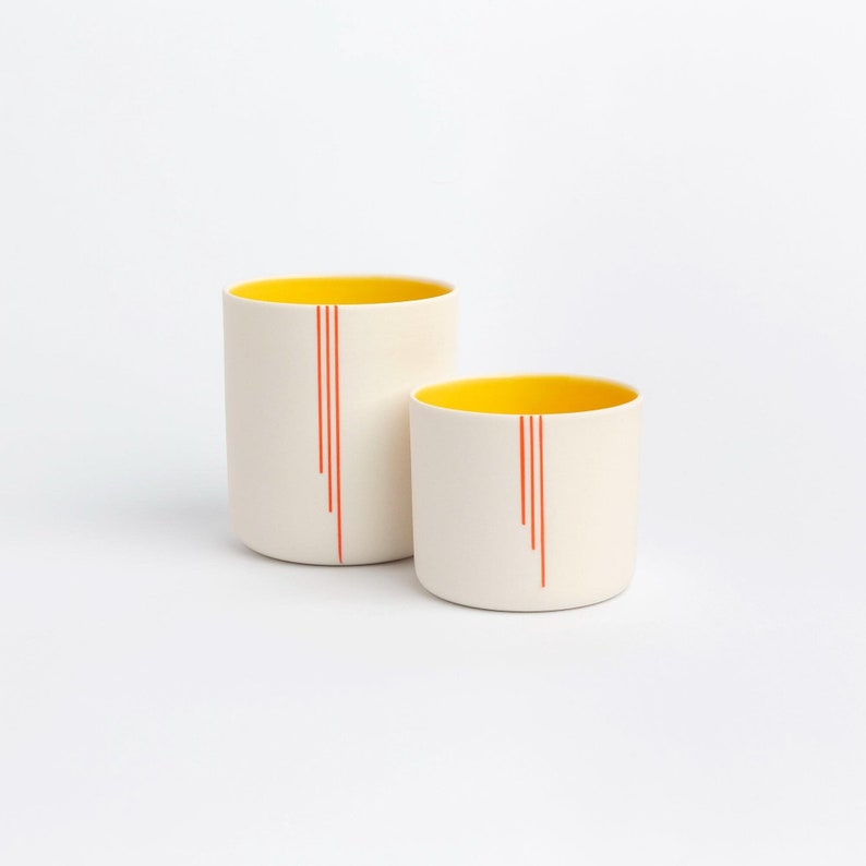 Peut inclure: Deux tasses en c&eacute;ramique avec des int&eacute;rieurs jaunes et des lignes rouges. Les tasses sont blanches avec une finition brillante.