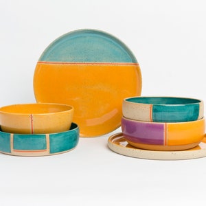 K&ouml;nnte beinhalten: Ein Set aus bunten Keramikschalen und Tellern mit geometrischen Mustern in den Farben T&uuml;rkis, Gelb, Orange und Lila. Die Schalen sind gestapelt und die Teller sind auf einer wei&szlig;en Oberfl&auml;che angeordnet.