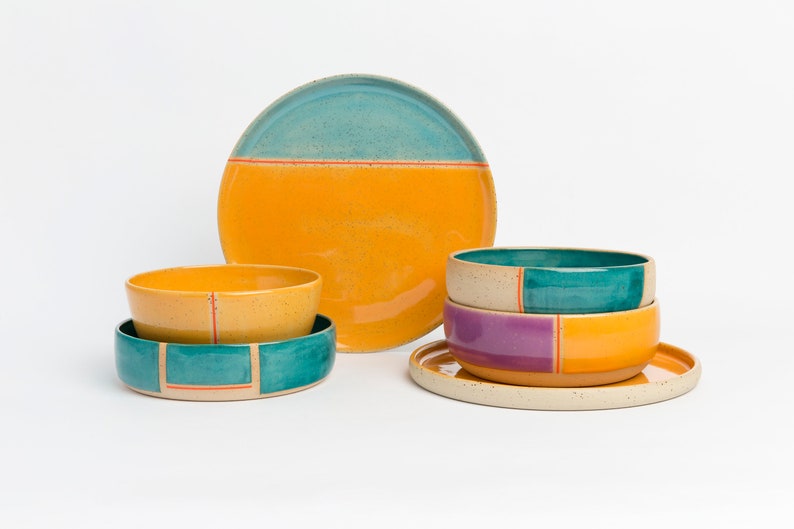 K&ouml;nnte beinhalten: Ein Set aus bunten Keramikschalen und Tellern mit geometrischen Mustern in den Farben T&uuml;rkis, Gelb, Orange und Lila. Die Schalen sind gestapelt und die Teller sind auf einer wei&szlig;en Oberfl&auml;che angeordnet.