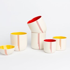 Peut inclure: Ensemble de cinq tasses en c&eacute;ramique avec des rayures rouges et jaunes. Les tasses sont empil&eacute;es les unes sur les autres, la tasse du haut &eacute;tant inclin&eacute;e sur le c&ocirc;t&eacute;. Les tasses sont toutes blanches avec des rayures rouges et jaunes.