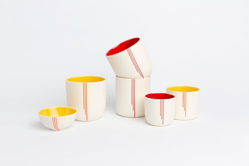 Peut inclure: Ensemble de cinq tasses en c&eacute;ramique avec des rayures rouges et jaunes. Les tasses sont empil&eacute;es les unes sur les autres, la tasse du haut &eacute;tant inclin&eacute;e sur le c&ocirc;t&eacute;. Les tasses sont toutes blanches avec des rayures rouges et jaunes.