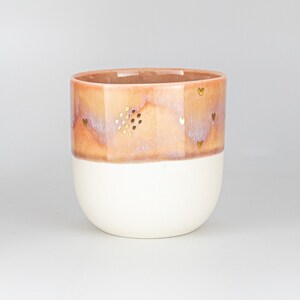 May include: A ceramic planter with a white base and a light pink top with a speckled glaze. The planter has a gold heart design.
