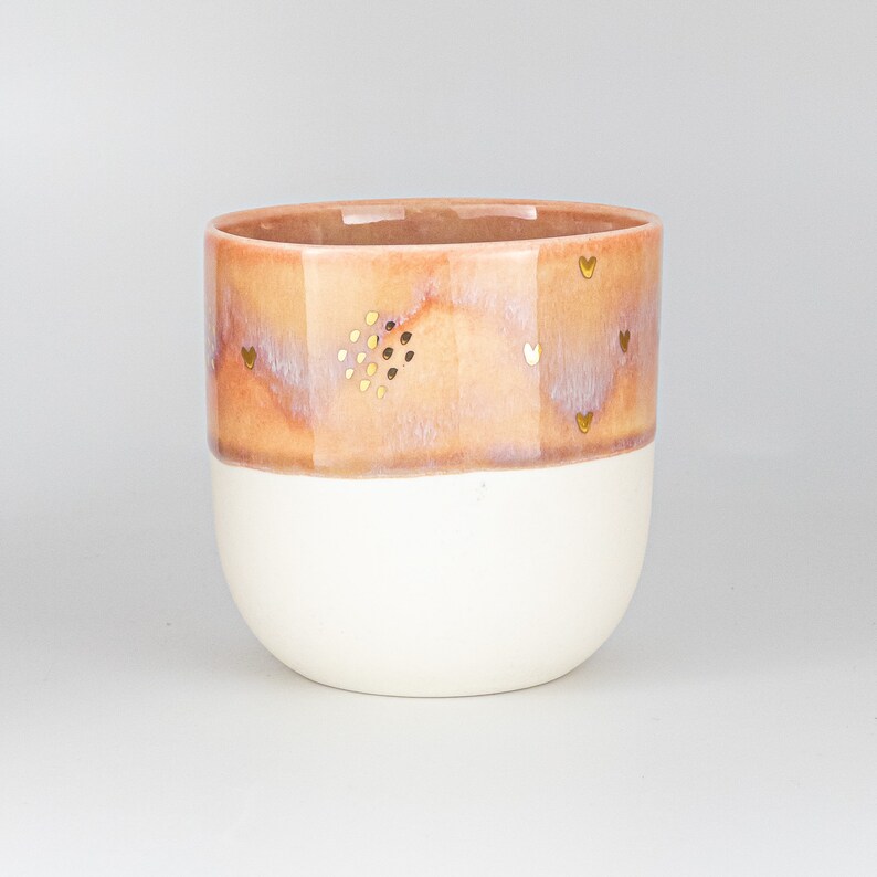 May include: A ceramic planter with a white base and a light pink top with a speckled glaze. The planter has a gold heart design.