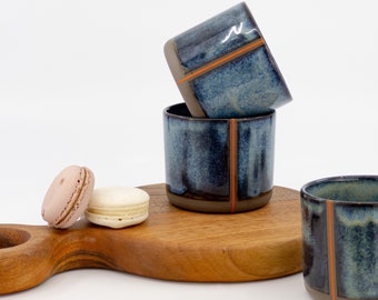 May include: Three blue ceramic mugs with orange stripes on a wooden cutting board with two macarons.