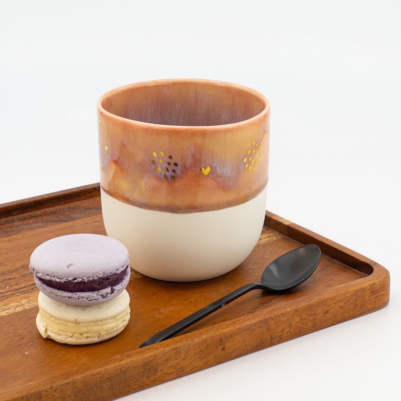 May include: A pink and white ceramic mug with gold accents and a heart design. The mug is sitting on a wooden tray with a purple and white macaron and a black spoon.
