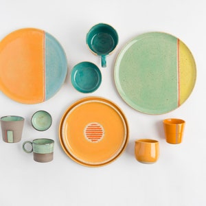 K&ouml;nnte beinhalten: Ein Set aus Keramiktellern und Tassen in verschiedenen Gr&uuml;nt&ouml;nen, Gelb und Orange. Die Teller haben ein schlichtes Design mit einer einzigen Linie, die die beiden Farben trennt. Die Tassen haben ein aufwendigeres Design mit einem Streifenmuster.