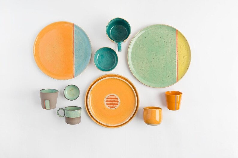 K&ouml;nnte beinhalten: Ein Set aus Keramiktellern und Tassen in verschiedenen Gr&uuml;nt&ouml;nen, Gelb und Orange. Die Teller haben ein schlichtes Design mit einer einzigen Linie, die die beiden Farben trennt. Die Tassen haben ein aufwendigeres Design mit einem Streifenmuster.