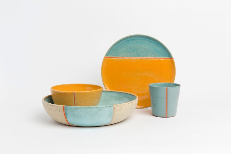 K&ouml;nnte beinhalten: Ein Set aus Keramikgeschirr in Blau, Gelb und Wei&szlig; mit orangefarbenen Akzenten. Das Set enth&auml;lt eine gro&szlig;e Sch&uuml;ssel, eine kleinere Sch&uuml;ssel, einen Teller und eine Tasse.