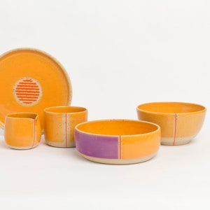 K&ouml;nnte beinhalten: Ein Set aus Keramikschalen und einem Krug in Orange- und Lilat&ouml;nen mit roten Akzenten. Die Schalen und der Krug haben eine gesprenkelte Glasur und sind handgefertigt.