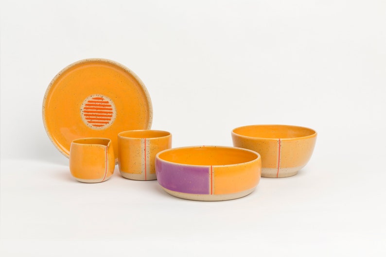 K&ouml;nnte beinhalten: Ein Set aus Keramikschalen und einem Krug in Orange- und Lilat&ouml;nen mit roten Akzenten. Die Schalen und der Krug haben eine gesprenkelte Glasur und sind handgefertigt.