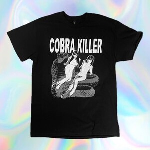 May include: Black t-shirt with the text "COBRA KILLER" in white block letters above a graphic of two women and a snake in white. The t-shirt is a crew neck style.