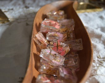 May include: A wooden tray holds several clear, rectangular resin pieces with pink and gold floral inclusions. The pieces have a cross design and are arranged in a wooden, curved tray. The tray sits on a white, textured surface.