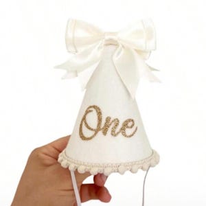 May include: A white party hat with a cream-coloured bow and the word "One" in gold glitter. The hat has a cream-coloured pom-pom trim and a string to secure it. A festive accessory for a first birthday celebration.