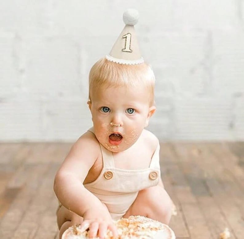 May include: A baby wearing a white felt birthday hat with a gold number one on it. The baby is sitting on a wooden floor and has cake on their face.