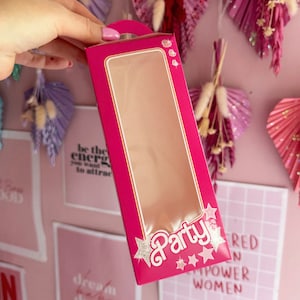May include: Pink cardboard box with a clear window, featuring the word "Party" in white with silver glitter stars. The box has a handle on the top.