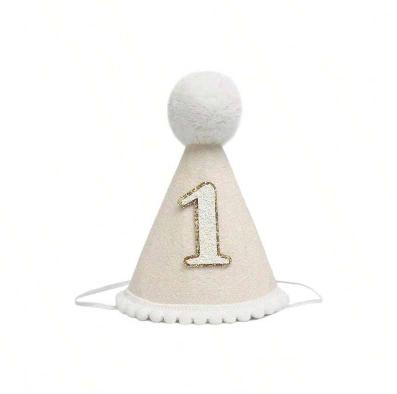 May include: A white and gold party hat with a fluffy white pom-pom on top and the number "1" in gold glitter.