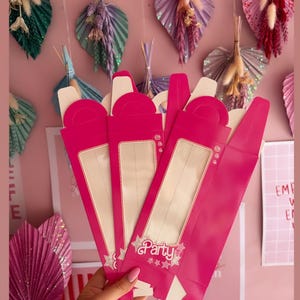 May include: Four pink party favor boxes with clear windows and a gold star design. The boxes have a handle on the top and say "Party" on the front.