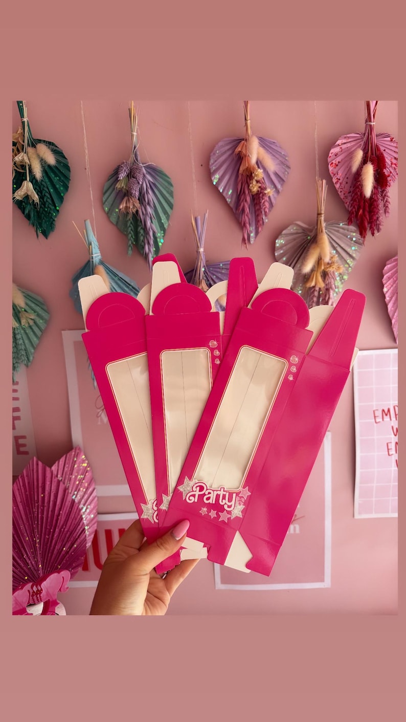 May include: Four pink party favor boxes with clear windows and a gold star design. The boxes have a handle on the top and say "Party" on the front.