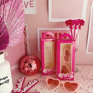 May include: Two pink boxes with clear windows, each containing a pair of pink heart-shaped sunglasses. The boxes have a white "Party" label with stars. The boxes are on a pink surface with pink glitter.