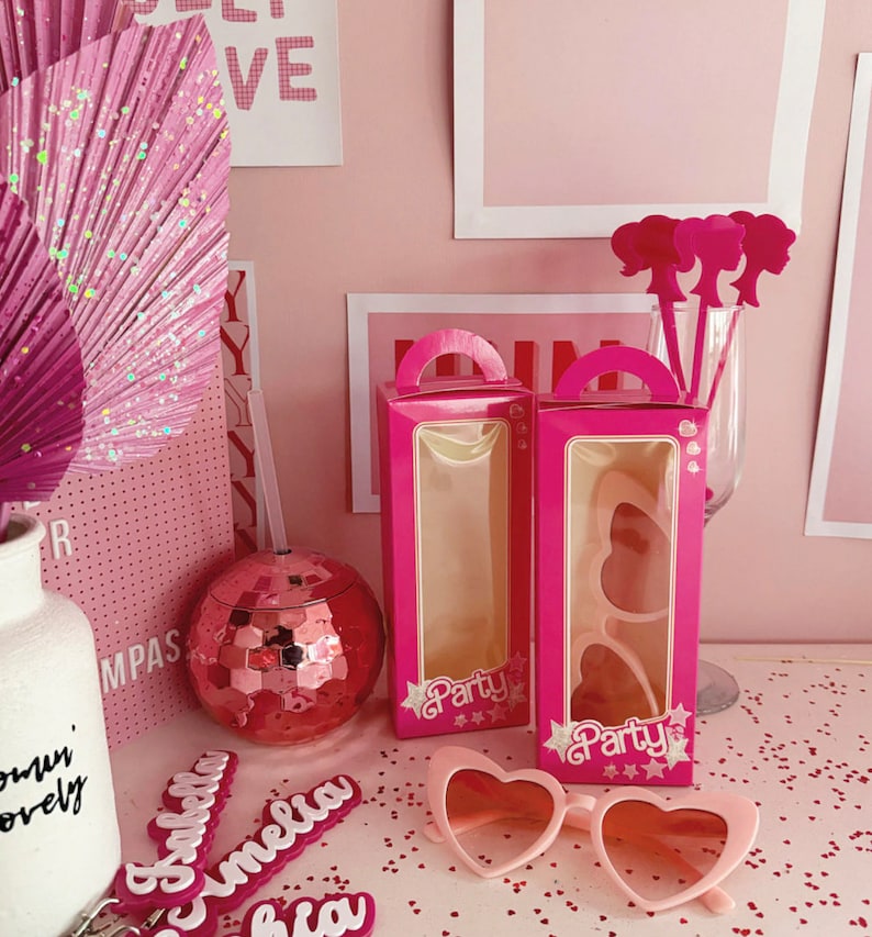 May include: Two pink boxes with clear windows, each containing a pair of pink heart-shaped sunglasses. The boxes have a white "Party" label with stars. The boxes are on a pink surface with pink glitter.