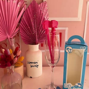 May include: A blue box with the text "Let's go Party" and a palm tree design. The box is open and there are blue star-shaped sunglasses inside. There are also pink flamingo-shaped drink stirrers in a clear glass.