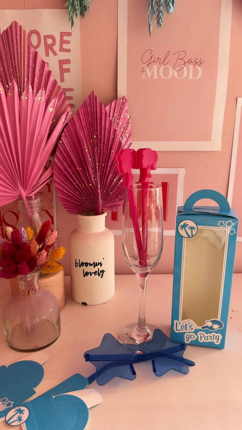 May include: A blue box with the text "Let's go Party" and a palm tree design. The box is open and there are blue star-shaped sunglasses inside. There are also pink flamingo-shaped drink stirrers in a clear glass.