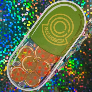 May include: A green and gold capsule-shaped sticker with the text "Capsule Corp." and a stylized "C" inside a circle. The sticker has a clear plastic overlay with a design of orange Dragon Balls with red stars.