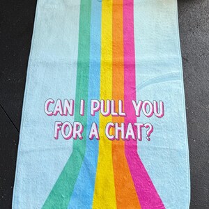 May include: A light blue gym towel with vertical rainbow stripes and the text "CAN I PULL YOU FOR A CHAT?". The towel is next to a black kettlebell and a weight plate. The word "WORK IT" is in the upper right corner.
