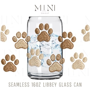 Puede incluir: Un vaso transparente en forma de lata con huellas de patas bordadas en tonos beige y marrón. El vaso contiene cubitos de hielo. El texto "MINI CRICUT" está en la parte superior y "SEAMLESS 16OZ LIBBEY GLASS CAN" en la inferior.